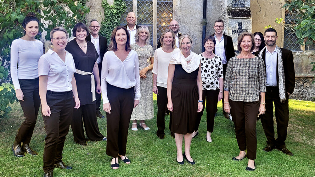 A portrait of a group of 15 people wearing a range of black and white smart-casual clothing standing on a lawn in front of an old church.