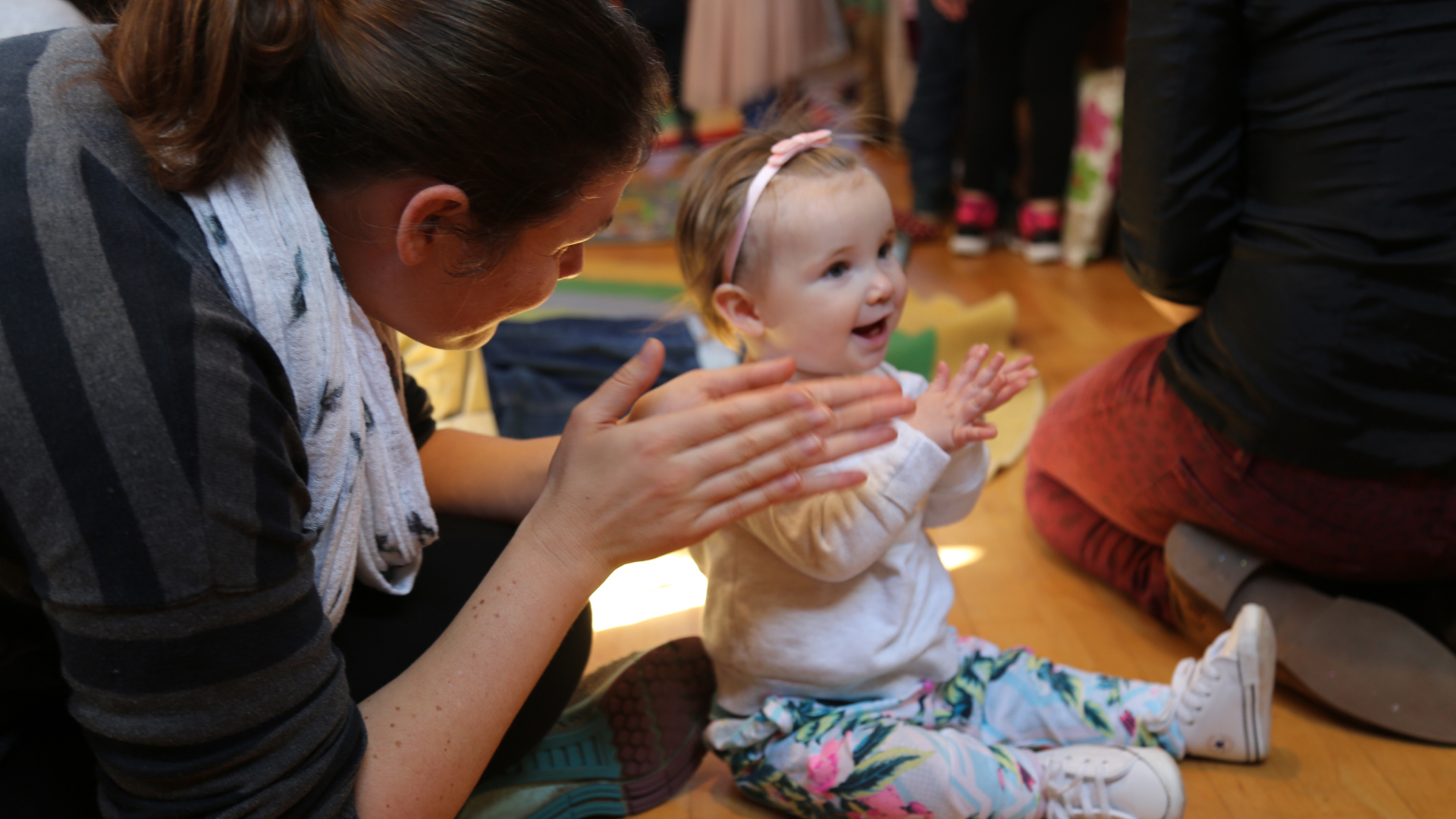 A baby and their parent sit on the floor looking happy and clapping.